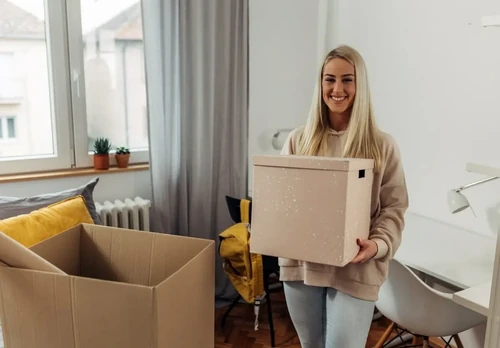 A college student smiles as she unpacks boxes of her items in her dorm room.