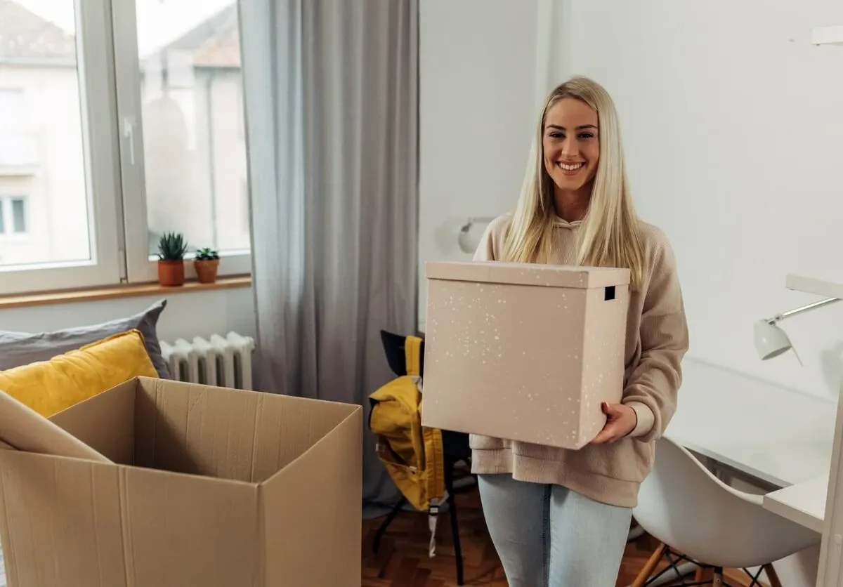 A college student smiles as she unpacks boxes of her items in her dorm room.
