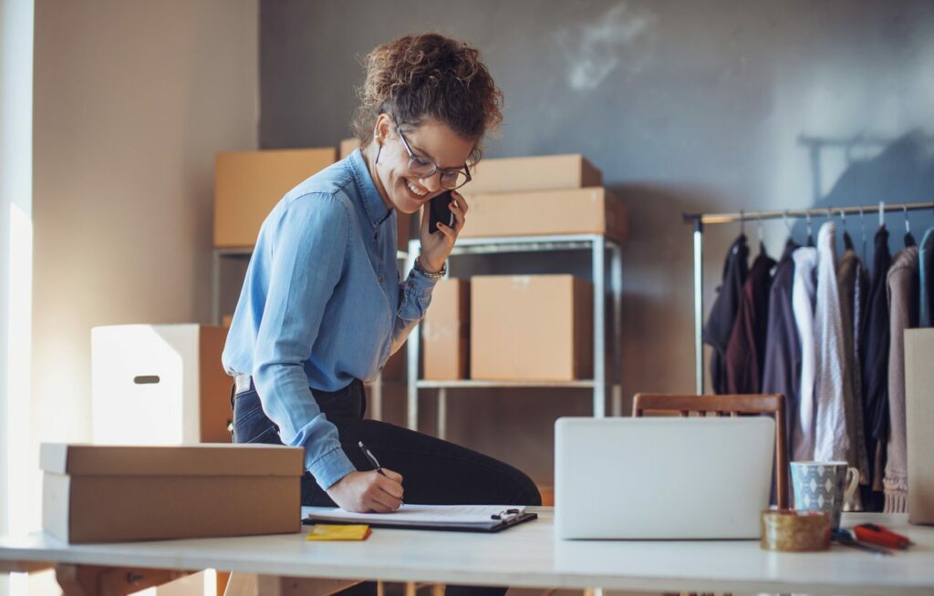A woman talking on a cell phone takes notes on a clipboard. Racks of clothing and boxes sit in the background.